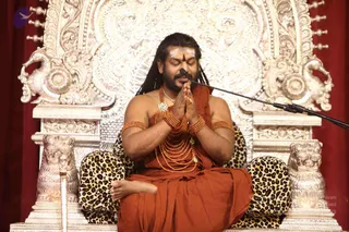 A seated man in saffron-orange robes sits on an ornate silver throne carved with lions and scrollwork, with leopard-print cushions, in front of a red curtain. He has long dreadlocked hair, a white tilaka with a red dot on his forehead, and wears multiple strands of rudraksha-style beaded mala, bangles, armlets and a large ceremonial pendant. His hands are pressed together in anjali and he is speaking into a microphone on a stand, eyes closed.