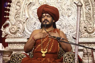 A photograph of a seated man in a saffron-orange turban and matching orange robes, wearing a long gold-toned necklace with a large pendant and rudraksha-style beads, with a small red mark on his forehead. He sits in front of an ornate silver-toned throne with carved floral and animal motifs, leopard-print cushions beneath him, and a silver staff beside him. He has his hands pressed to his chest while speaking into a microphone on a stand. A white cup sits on a side table at left.