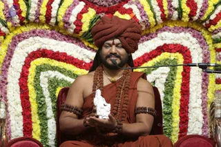 A man seated on a red throne-style chair in front of a large floral backdrop of concentric arcs of roses, marigolds and white flowers in red, yellow, green and white. He wears a rust-orange turban with a vermilion tilak, an orange-saffron cloth draped over his shoulders, multiple rudraksha malas, a gold chain, and rudraksha armlets. He smiles and holds a small white marble figurine of a seated sage in both hands, with a microphone stand in front of him. Swamiji (probable) appearance cues include the turban, saffron clothing, rudraksha malas and gold chain, while he is seated and presenting the statue.