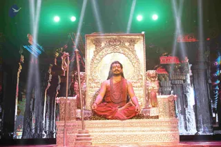 A photograph of a seated figure in meditation posture on an ornate gilded throne on a stage. The man wears long dark hair, an orange-red cloth wrap over bare shoulders, a long rudraksha mala, a gold necklace and a red-orange tilak on his forehead. He sits with hands in a meditation mudra before a large gold arched backdrop flanked by two golden lion figures and a leopard-print seat cover. A golden staff stands to his left. Green and white stage lights beam down behind him onto a set of painted temple pillars in red and gold. a person, seated in meditation on the throne, with his ornate saffron attire, rudraksha mala and gold necklace; one physical person visible.