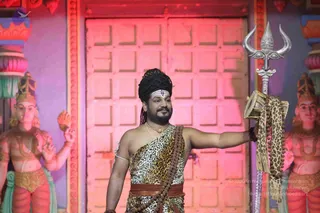 A man stands in front of a pink-lit wooden door and two painted deity statues with crowns and ornaments. He wears a black turban, a white tilak across his forehead, a leopard-print cloth wrapped over his torso, a rudraksha mala across his chest and a gold chain. He holds a large silver trident in his right hand, its shaft wrapped with a leopard-print cloth, while his left arm is extended to the side. The background shows red and purple pillars and a decorative carved panel.