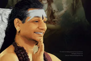 A close-up photo of a smiling man with long dark hair swept back, wearing a white horizontal tilak with a dark central dot on his forehead, gold earrings, a large rudraksha mala of dark beads, and a bare shoulder with a reddish-brown cloth. His right hand is raised to his chin in a gesture, and a gold ornament rests on his finger. Behind him is a painted image of a deity with multiple arms and a glowing dark background. Swamiji (probable): long swept-back hair, rudraksha mala and ceremonial gold ornaments are visible cues, presented as a satsang speaker.