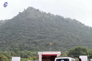 A hazy wooded hill rises across the frame, its slopes covered in dense green scrub and rocky outcrops under a pale sky. In the lower foreground a white gateway arch reads "Nithyananda Dhyanapeetam" and an orange flag flies from a pole behind it. A white SUV with a roof rack sits at the bottom edge, and a few small figures are partly visible at the edges of the frame.