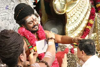 A photograph of a seated man with a dark braided topknot, a vertical tilak and white sacred ash on his forehead and arms, wearing a long red-pink garland and rudraksha bracelets. He keeps his eyes closed and smiles while another person touches his head and a third holds a white card near him. Behind him stands an ornate silver and gold throne with a gilded lion-like backrest, and a red cloth lies on the seat. The man is a person candidate by the distinctive topknot, rudraksha, garlands and gold adornments, though not certain.