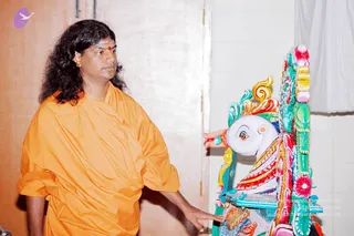 A man with long, shoulder-length dark hair and a red bindi on his forehead stands in an ashram room wearing a loose saffron-orange robe. He rests one hand on a large brightly painted wooden figure with a white face, red and green ornamentation and pink floral carvings, appearing to examine or touch the figure. Pale walls and a white curtain form the background. Appearance-based label: Swamiji (probable), wearing saffron robe with long hair and a bindi, interacting with the painted ratha figure.