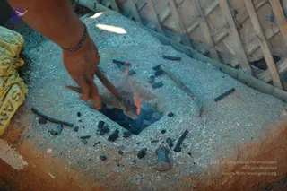 A close photograph of a hand using a pair of metal tongs to work over a small charcoal fire pit lined with glowing embers and scattered black charcoal pieces. The pit sits on a rough earthen or clay platform dusted with grey ash. A woven bamboo wall is in the background, and a yellow cloth lies at the left edge. A metal hammer or tool head rests near the bottom of the frame. The hand wears a braided wrist cord. No people's faces are visible, and no a person candidate is present.