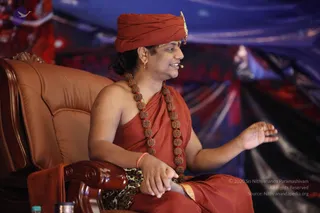 A man seated in a brown leather armchair with carved wooden armrests speaks during a talk, one hand open in a gesturing pose. He wears a rust-orange turban with a small gold ornament, an orange-red shawl draped over bare shoulders and a long mala of large rudraksha beads. A small microphone is clipped near his mouth, and a dark leopard-print cloth lies beneath him. The background shows a blue and red draped stage backdrop. The person matches appearance cues for a person, sitting as a speaker on stage.