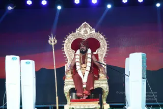 A man in saffron-red robes and a matching red turban sits on an ornate gilded throne with a red cushioned back, a golden trident leaning beside him on the left. A long white and red garland hangs around his neck, with a gold pendant and beaded necklaces on his chest, and he rests his hands on the armrests while speaking into a microphone stand. Two white tower speakers flank the stage, which has a dark red-to-orange sunset backdrop with blue stage lights above. The appearance-based label is Swamiji (probable), given the saffron-red clothing, red turban, ornate gold necklaces and the throne setting, though this is an appearance-based candidate only.