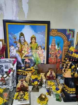 A home shrine with no physical people visible. A large framed print of Shiva seated with Parvati, Ganesha and young Murugan on a mountain backdrop, with a Shiva lingam and a white bull at left. Beside it stands a painted panel of a four-armed goddess holding a sugarcane stalk and a mirror-like object within a golden arch. Flower garlands in pink, white and yellow hang on the frames, and yellow flowers decorate the altar. A black sculpted deity figure with many arms stands at right. Small seated portraits of a robed teacher with a garland sit on the altar, along with ivory elephants, a brass kalasha-like vessel and a framed black-and-white photograph.
