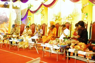 A seated panel on a red carpet under a yellow and purple draped stage with white flower garlands at a book release event. Centre, a man in an orange robe and orange turban sits with palms pressed together in greeting, a microphone before him. To his left, a woman in a white and gold sari and an elderly man in a white shirt and dark cap sit with hands folded. To his right, a man in a white shirt with glasses and two men in dark shirts sit in red-cushioned carved chairs. Low glass tables hold fruit bowls and name cards. A banner with a portrait stands at left.