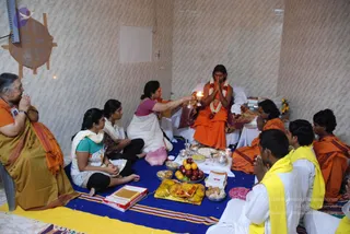 A seated man in orange robes and a white flower garland receives a camphor aarti from a woman in a white sari with a magenta blouse, during a puja on a blue cloth with yellow edging. A row of devotees sits around him: a woman in a mustard sari with an orange shawl presses her palms together at left, two young women sit in white and teal salwar dress, and young men in white dhotis with yellow shawls sit at right, one with hands folded. Fruit, sweets, flower petals, steel vessels and boxed sweets are arranged on the cloth in front. The wall is pale tiled with a round wooden plaque at left.