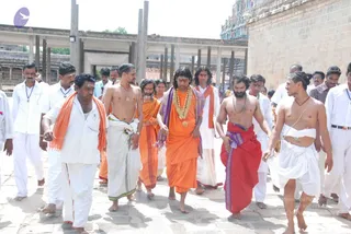 A procession of men walks barefoot across a stone-paved temple courtyard beside a stone wall and a colourful gopuram tower. At centre, a man in saffron-orange robes wears a long orange-and-purple shawl, a large marigold garland and a ceremonial gold pendant, with long swept-back dark hair and a painted forehead mark; he has distinctive long hair, saffron clothing and a garland, so this is an appearance-based probable Swamiji candidate in the procession. Around him, men in white dhotis and shirts, some in orange shawls, and bare-chested men with rudraksha-style neck beads and red or orange cloths walk alongside. Several men wear orange scarves and rudraksha malas. About fifteen people are visible.