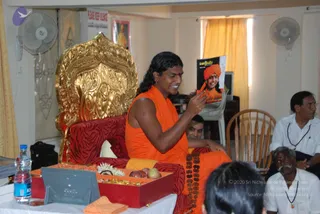 A seated man in an orange-saffron garment with long dark swept-back hair sits on an ornate golden throne upholstered in red and white. He holds up a magazine cover showing a smiling man in an orange turban and orange clothing, while smiling. A red tray with fruit and flowers sits on the table in front, beside a bottle of water. Two men in white shirts with rudraksha malas sit at right, and a person's head is visible at lower right. Fans and curtains are in the background.