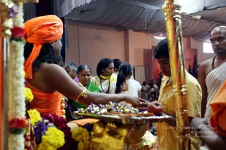 Photograph of a ritual scene inside a hall with a white draped canopy. In the left foreground a person in saffron-orange clothing with a saffron-orange turban reaches toward a brass tray of flower petals and offerings, beside a golden staff and garlands of white jasmine and red and yellow flowers. Behind, a woman in a green sari and several women and girls in white and yellow attire watch closely. On the right a bald man with a rudraksha-style necklace and a white dhoti, and a young boy in yellow kurta hold items near the tray. The a person candidate in saffron with a turban is visible from behind and to the side, so identity is uncertain.