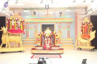 A seated man in a red garment sits on a gold-and-red cushioned throne (peetha) at the centre of a decorated stage, wearing a white turban-like headwrap and a long white flower garland. The throne is framed by an arch of garlands in red, green, yellow and purple. Flanking him are two standing attendants in orange-and-gold clothing holding staffs. On the left, a golden bull (nandi-style vahana) carries a garlanded pair of deities under a red canopy; on the right, a golden peacock-like vahana carries a garlanded standing figure in orange. A small red rug lies in front, and a camera and monitor are visible in the foreground. Pale blue temple-style columns and gold arches decorate the backdrop.