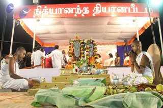 Night outdoor ceremony under a white canopy with orange and gold valances. Two shaved-headed priests in white dhotis sit on the ground in front of a raised gilded platform. The priest on the left plays a small hand instrument. The priest on the right sits with palms pressed together in anjali beside a bronze and silver kalasha. Two deity murti are dressed in orange and red garments, garlanded with multicoloured flower malas, and stand before a bowl of offerings with banana leaves and bananas and mangoes. Devotees in white and colourful saris stand behind the platform, and stage lights hang above. A Tamil banner reads Nithyananda Dhyanapeetam. Event satsang or puja.