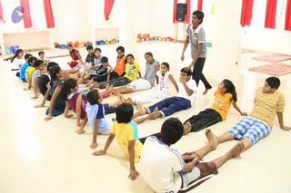 A photograph of a bright, cream-walled hall with red curtains where a group of children sit on the floor in two facing rows, many with legs stretched forward. A standing teen in a grey polo shirt walks between them near a black speaker on a stand. Children wear white, yellow, orange, blue and striped clothes. Red and pink mats lie on the floor and coloured boxes are stacked along the back wall. The setting suggests a children's class or activity session, not a specific ritual.