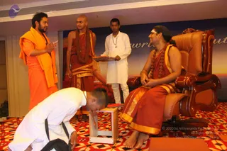 A photograph from a stage event with a red, orange and yellow patterned carpet and a backdrop with partial text reading "rewri" and "ut". A seated man in a saffron-and-gold dhoti with a rudraksha mala sits in a large brown leather throne-style chair, smiling, barefoot. A standing man in an orange robe and orange shawl with a tilak gestures with his hands in conversation. A shaved-headed man in red-brown robes with a long rudraksha mala stands beside them. A man in white kurta holds a tray. In the lower left, a man in white kurta bows low with his head near a small white stool with a red cushion.