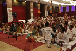 A large indoor hall with red carpeting and white floor cloths where a seated audience fills most of the frame, many in white, saffron and orange clothing with orange garlands and saffron shawls. In the foreground a small boy in a white kurta walks toward a seated man in white clothes with an orange garland, while a television camera on a tripod and a second camera stand at left. Hanging colourful decorative tassels, chandeliers and purple lit panels decorate the back wall, and a small figure in orange stands near the rear stage area. Attendees sit in rows on the floor for a satsang-style session.