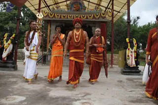Five men in ceremonial dress walk across a paved courtyard in front of a shrine with a deity murti in an arched niche and a yellow-and-green canopy overhead. The central man wears a red-maroon turban, a gold ornamental necklace, a rudraksha mala and maroon robes edged in gold, and the man to his left wears orange robes with a rudraksha mala. A man on the far left in white robes holds a golden trident aloft, and a bald man beside the central figure in maroon robes holds an orange cloth bundle. Garlanded black stone statues stand on pedestals at both sides. Trees and a stone wall form the background.