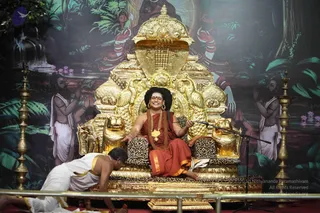 A seated man with long swept-back dark hair, a red tilak and a red-saffron robe with a gold border sits on an ornate gilded throne with lion-head armrests and leopard-print cushions, smiling and gesturing with one hand. A gold pendant and a beaded mala hang at his chest and large gold earrings are visible. Before him, a man in white dhoti with a gold border bows low at the base of the throne. Behind the throne, a painted backdrop shows a serpent-hooded deity form and figures with folded hands beside a lotus pond, with tall brass lamps flanking the stage. A microphone stand is in front of the throne.