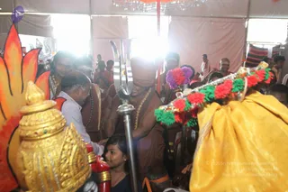 Inside a bright white tent, a bald man in a rust-brown robe with a long gold-toned necklace stands beside a tall silver trident (trishul) held upright. Around him, several people in brown and red-brown robes and white clothing gather among devotees. A girl in a dark top stands near a man in a light shirt who bends forward and touches a ceremonial object. In the foreground, a gold-decorated kalasha (ritual pot) sits beside a red-and-gold object, and a yellow cloth draped over an item is trimmed with red, green and purple pompoms. Behind them, a red-and-orange flame-shaped backdrop and hanging crystal chandelier are visible.