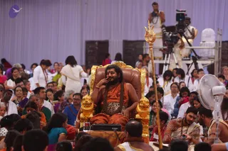 A large indoor hall with a pale lavender draped backdrop and many seated devotees in white and colourful saris and kurtas. At centre, a man with long dark hair and a full beard sits on an ornate gold throne with carved lion armrests, wearing an orange-saffron robe and a long rudraksha-style mala, with a leopard-print cloth across his lap; he rests a finger near his mouth. A tall golden trident (trishul) stands beside the throne. Behind, a cameraman operates a video camera on a tripod, and a person in white dhoti stands on a raised platform. A white electric fan sits at lower right. Appearance-based candidate: Swamiji (probable), seated on the throne and wearing saffron robes, long hair and a rudraksha mala.