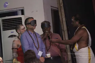 A stage event where two men wear black blindfolds. The man in the centre-left wears a light blue button shirt and a rudraksha mala, and the man beside him wears a pink shirt, a rudraksha mala and an ID badge on a lanyard while holding a microphone. On the right, a shirtless priest in a white dhoti with a yellow border, a rudraksha mala and a white forehead mark holds the microphone toward them. A shaved-headed young man in a red garment with white body markings stands at the left edge.