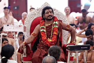 A man seated on an ornate silver throne with red velvet cushions, wearing a rust-brown garment over the shoulders, a dense garland of red and white flowers, gold chain necklaces, a gold-bordered orange wrap, and beaded gold armlets. He has a vertical tilak on his forehead and keeps his eyes closed with a calm expression, one arm resting on the armrest. Around him seated devotees, several bare-chested, some in white, surround the scene. A Yamaha keyboard sits on a silver stand to the right.