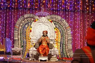 A seated figure in orange-saffron draped clothing and a red-orange turban sits on an ornate silver throne with a large embroidered arch of gold and jewel-toned panels behind it, topped by a silver canopy. He wears a multi-strand red-and-gold beaded necklace and bangles, raises his hands in front of his chest, and speaks into a microphone on a stand. Garlands of yellow and red flowers drape the throne arch, and strings of multicoloured fairy lights hang across the crimson backdrop. Blurred red-clad figures are visible at the right edge.