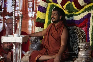 A seated man in an orange-rust saffron robe sits on a leopard-print cushioned throne with silver lion-head armrests, smiling and resting one hand on a silver lion figure. He has long dark hair, a red tilak on his forehead and a beard. Behind him is a wall of colourful garlands in red, purple, yellow, green and white. At left, a silver stand holds a small white cup, and two seated devotees with folded hands are partly visible. The man is an appearance-based probable Swamiji candidate: long swept-back hair and a saffron robe, with the page indicating a satsang setting.