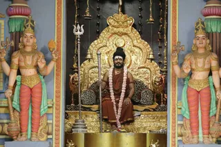 A seated man in saffron-orange robes sits on an ornate gold throne with leopard-print and tiger-skin cushions, holding a silver trident-topped staff. He wears a rudraksha mala, a fresh pink and white flower garland, a dark turban-style headwrap and a gold necklace. Flanking him are two painted, life-size deity murti figures with crowns, four arms, gold jewellery and green sashes, holding ritual objects. A golden canopy with hanging bells and a gold kalasha-like finial frames the shrine, with a gold base below. Swamiji (probable) is seated on the peetha as the central figure, with long swept-back hair and saffron/orange clothing and rudraksha cues visible.