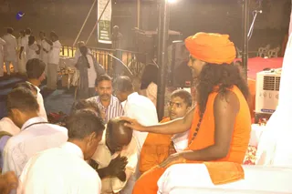 Night outdoor event with a crowd of men in white shirts and dhotis. On the right, a figure in an orange turban, long swept-back dark hair, orange robe and rudraksha mala sits on a raised white seat and rests a hand on the head of a seated devotee in a white shirt with bowed head and folded hands. Another man in an orange robe sits beside him. A pedestal fan, a white-draped table with flowers, a loudspeaker and a green banner stand nearby.