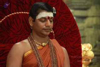 A man seated with eyes closed on a red throne upholstered in a dark red tiger-striped fabric with gold cord trim, a gold ornament at lower right. He wears an orange-saffron robe with a gold border, multiple strands of rudraksha beads, a gold pendant necklace, hoop earrings, a white horizontal forehead band with a red dot at the centre, and a long swept-back hairstyle with a decorative hair ornament. His hands are not visible. The background is a blurred stone wall.