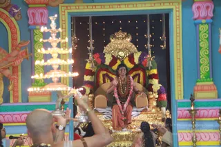 A photograph of a seated figure on a gilded throne with a crown-shaped golden canopy, draped in a garland of red, yellow, and purple flowers over an orange-brown cloth, with rudraksha beads and gold necklaces, hands raised in greeting. A tiered brass aarti lamp with many burning flames is held in front of him by a bald man in white clothes at the lower left, and a few other devotees sit at the lower right. The throne is set within a blue and pink temple-style backdrop with painted pillars and a hanging string of lights. Swamiji (probable): the seated figure wears long swept-back hair, rudraksha mala and gold ceremonial necklaces, and orange-saffron clothing, consistent with the page's account of a satsang on the throne.