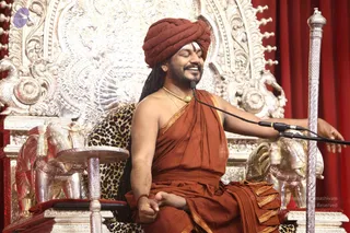 A man seated on an ornate silver throne with carved floral and animal motifs, eyes closed and smiling broadly during a discourse. He wears a rust-orange turban with a white forehead mark, an orange shawl draped over one shoulder and a saffron-orange wrap, a gold necklace, and a red thread on his wrist. A microphone stands before him, a silver staff rises at right, and silver elephant figures flank the throne against red drapes. A leopard-print cloth covers the seat.
