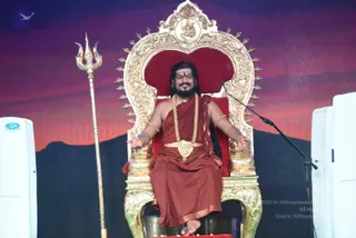 A man seated on an ornate gold-and-red throne on a stage, arms spread open, wearing a rust-red draped garment, a rudraksha-style beaded mala and a large ceremonial gold necklace, with gold bangles on his arms and a red tilak on his forehead. A golden trident stands to his left, a microphone stands to his right, and a sunset-coloured backdrop with dark hills sits behind him. Two white cooling units flank the stage. a person is seated here as the central figure, with the throne and trident marking the ceremonial setting.