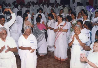 Photograph of a large group of devotees dancing and clapping in a hall with a brown tiled floor, most dressed in white. Foreground: an older woman in a white sari with a red bindi and glasses, hands pressed together in namaste; beside her a woman in a white sari with red and orange border, clapping; a man in white dhoti at left with glasses, clapping. Behind them a woman in a grey-and-black sari and pink blouse with hands together, and several people raising arms. Name badges are visible on some white-clad participants. No central figure in saffron or crown is clearly visible, so no candidate is identified. Background shows more participants clapping and swaying.