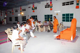 A hall with whitewashed walls, a metal roof and ceiling fans hangs colourful striped cloth banners from the rafters. A man in saffron-orange robes stands on a rug at right, speaking with a hand raised and a microphone on a stand beside him. Beside him sits a large orange-draped chair, a peetha-style seat with a red-edged rug beneath it. Around 15 seated men in white shirts, dhotis and trousers listen in plastic chairs, some holding notebooks or bags. A woman in orange and a person in white stand near a tripod-mounted camera at the back, and a few more people move near the left wall. Water bottles sit on a small table near the seat.