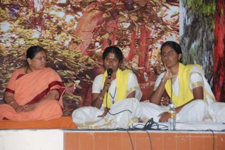 Three women sit on a raised stage in front of a large printed backdrop of a forest with red-brown tree trunks and green leaves. The woman on the left wears an orange saree with a maroon border and rimless glasses, seated on an orange cloth. The woman in the centre wears a white saree with a yellow shawl and a rudraksha-style mala, holding a black microphone while speaking. The woman on the right wears a white saree with a yellow shawl and a beaded necklace, one hand resting on her cheek. A water bottle sits on the stage edge.