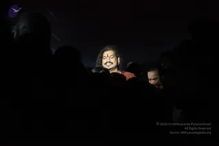 A dark night photograph of a crowd with a single man lit by a spotlight at the centre. He has long, swept-back dark hair, a red tilak on his forehead, a moustache, a gold chain and a red-orange shawl, and he smiles with eyes lowered. Silhouetted heads surround him. To the right, a bearded man with white horizontal ash lines on his forehead looks down. Probable Swamiji shown with long hair, orange-red wrap and gold necklace cues, presented as a person in the crowd; the surrounding figures are devotees or attendees in darkness.