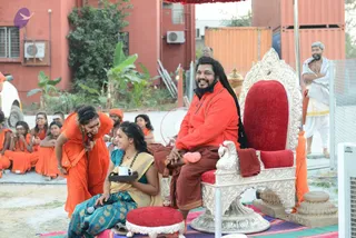 Outdoor courtyard session. A bearded man with long dark hair wearing an orange-red top and rust-brown dhoti sits smiling on an ornate silver throne with red velvet cushions, a swan-carved armrest visible. Beside him a woman in a teal blue and gold saree holds a small white cup on a black tray and a microphone. In front, a woman in orange robes bends forward smiling. Behind them a group of young people in orange clothes sit on the ground, and a man in white dhoti and shawl stands at right near a chain-link fence. Red brick building, banana plants and shipping containers in the background, with a red umbrella overhead and a red-cushioned stool at lower left.