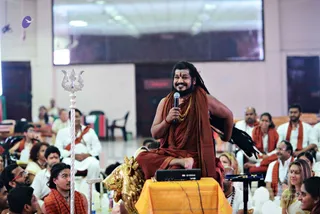 A seated man in an orange-red saffron robe, with long swept-back dark hair, a beard, a red tilak on his forehead and a heavy gold beaded necklace, holds a handheld microphone and smiles while speaking. He sits on a raised orange-draped platform behind a black keyboard, with a golden lion-like figure beside him and a silver trident standing to his left. Around him an audience of mostly white-clad devotees, several wearing orange or saffron shawls, sits in rows of white chairs under a large screen in a hall. Swamiji (probable) is shown at the centre as the speaker in saffron, with cues of long hair, saffron robe and gold necklace.