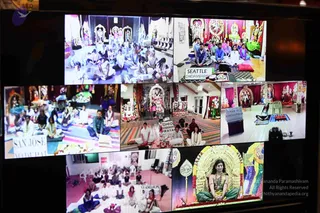 A photograph of a wall-mounted video display showing a multi-panel collage of live video feeds from several centres. Panels show groups of people seated on cushions and mats in front of decorated altars with flowers, garlands and deity images. Labels read SEATTLE CHIDAMBARAM, SAN JOSE MADURAI, OHIO PRAYAG, PHOENIX KANCHEEPURAM and GUADELOUPE. In the lower right panel a figure with long dark hair wears a green and gold garment and sits on a gold-framed throne before a golden deity arch decorated with marigold garlands. Physical people visible in the room photographing the screen are not counted.