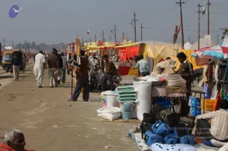 A dusty dirt road lined with open-air market stalls under a hazy sky, with utility poles and yellow and red tarpaulin awnings. Vendors stand behind piles of white sacks, stacked plastic-wrapped packets, and blue and black travel bags, while shoppers and passers-by in white kurtas and warm jackets walk along the road. A seated elderly man in an orange garment sits at the lower left, and a three-wheeled auto-rickshaw is visible at the left edge. Strings of colourful garlands or threads hang from a stall at left. The scene appears to be a busy temporary bazaar.
