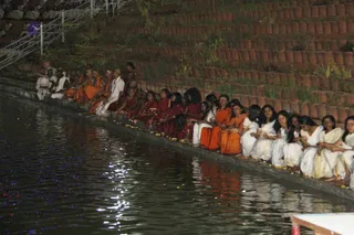Night photograph of a long row of people seated on the stone edge of a reflective water tank, with dark water in the foreground and terraced terracotta-tiled steps rising behind them. Many seated figures wear saffron and orange robes, some in deep red, while others wear white dhotis and white shawls, several with shaved heads. Some hold small lamps or flower offerings, with marigold petals scattered along the water's edge. A figure in white at the far left appears to sit higher on a platform, but the scene does not clearly show who is leading the ceremony. Warm lights reflect on the water.