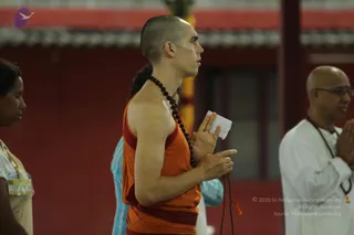 Photograph of a shaven-headed man in an orange sleeveless robe with a red waistband and a long rudraksha mala hanging to his waist, standing in profile with hands joined in anjali mudra and a folded white cloth held between the palms. Beside him at left a woman in a light beige top with a name badge stands partly visible, and at right an older bald man with glasses wears a white kurta and a rudraksha mala with hands folded in prayer. A woman in a blue garment is partly behind the central figure. The background is a red wall and pillar.