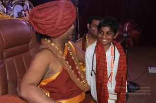 A seated figure in a red-orange turban, orange-gold garment and a heavy beaded gold necklace sits on a brown leather chair, smiling toward a young boy. The boy wears a white kurta, a red scarf printed with script, a rudraksha mala and an orange Kumbh Mela lanyard badge. Behind them a man in an orange shawl and gold necklace smiles, and a red oscillating fan stands at right. A gold-ornamented shrine with a deity figure is visible at the upper left. The seated figure shows distinctive ceremonial cues, a turban, ornate gold necklace and saffron clothing, so this is a probable appearance-based match for a person in person.