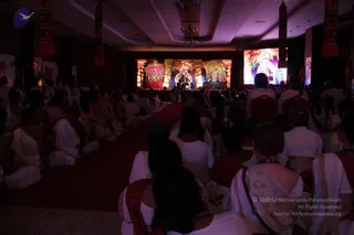A dimly lit hall with an audience of many devotees seated on the floor facing a stage. Most attendees wear white clothing with red or maroon shawls draped over their shoulders, and some are seated on wooden chairs. The stage is framed by decorative arches and lit by warm lamps, with elaborate deity murti or decorated figures displayed in the centre and at each side. A large screen on the right shows a colourful image. Red carpet runs down the centre aisle, and petals are scattered on the floor. Hanging saffron and red fabric banners line the upper walls.