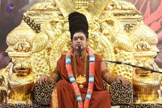 A seated man in an ornate golden throne with lion-head armrests and a golden peacock-like backrest sits before a microphone. He wears a rust-orange robe, a long gold chain with a large gold pendant, rudraksha beads, and a multicoloured garland of pink, white and blue flowers. His long black hair is piled high on his head, and he has a red bindi and white markings on his forehead. Leopard-print cushions rest on the throne's arms.